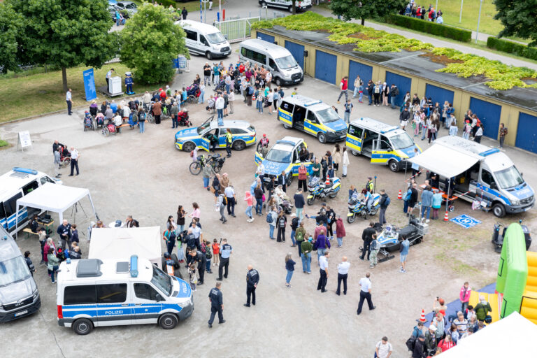 Aerial view of a public safety event with Polizei vans, a patrol car, tents, and a crowd of adults and children in a parking lot area.