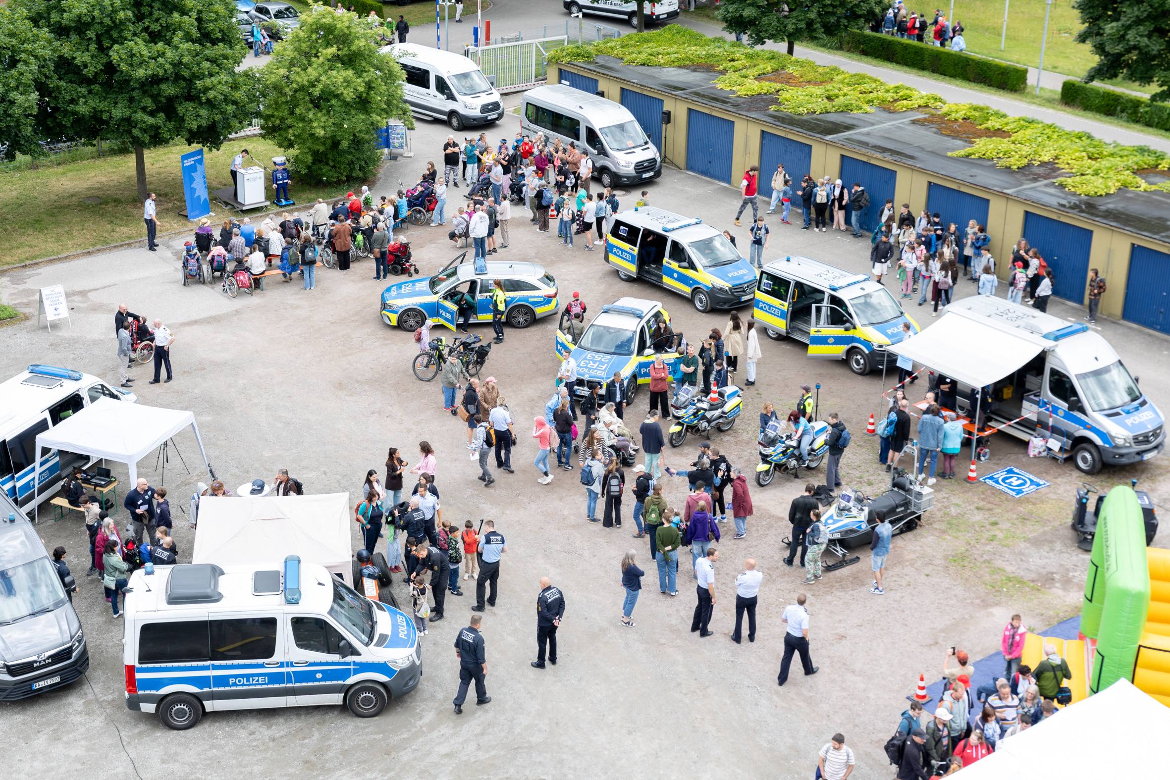 Aerial view of a public safety event with Polizei vans, a patrol car, tents, and a crowd of adults and children in a parking lot area.