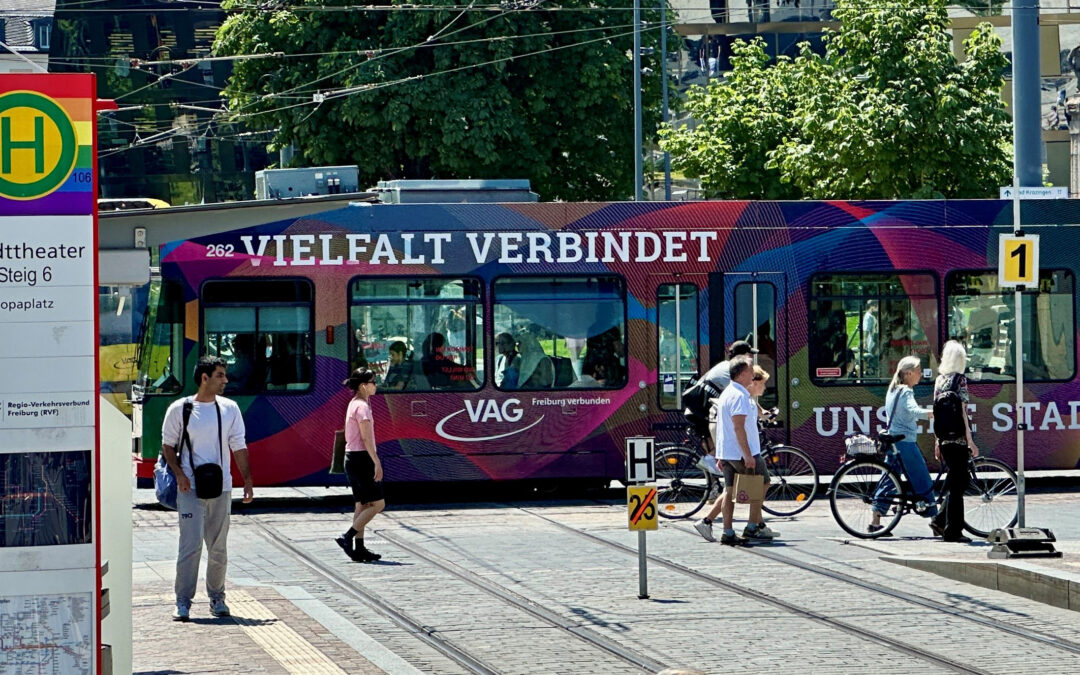 Multicolored tram with 'VIELFALT VERBINDET' lettering at a sunny city stop, pedestrians and cyclists nearby.