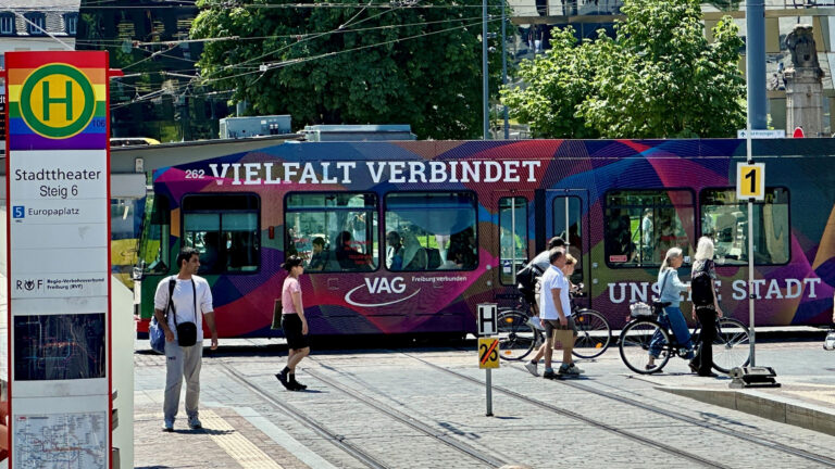 Multicolored tram with 'VIELFALT VERBINDET' lettering at a sunny city stop, pedestrians and cyclists nearby.