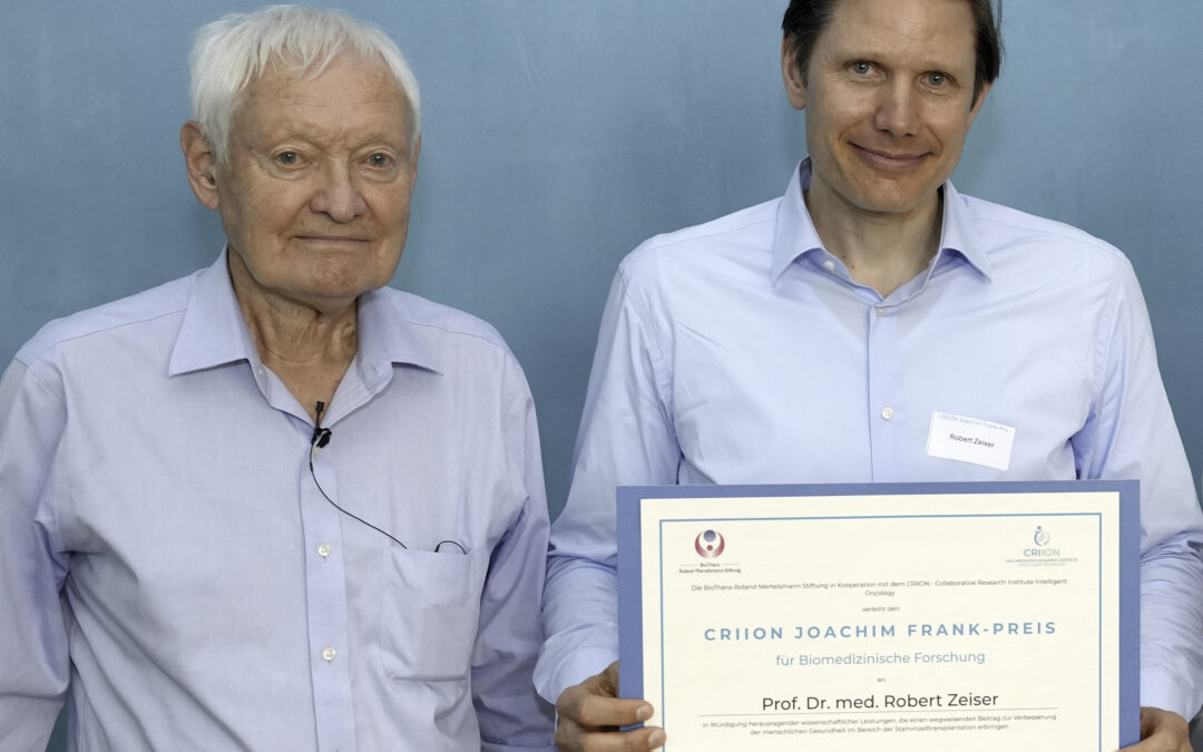 Two men in light blue shirts pose with a large framed certificate against a blue wall, suggesting an award ceremony.