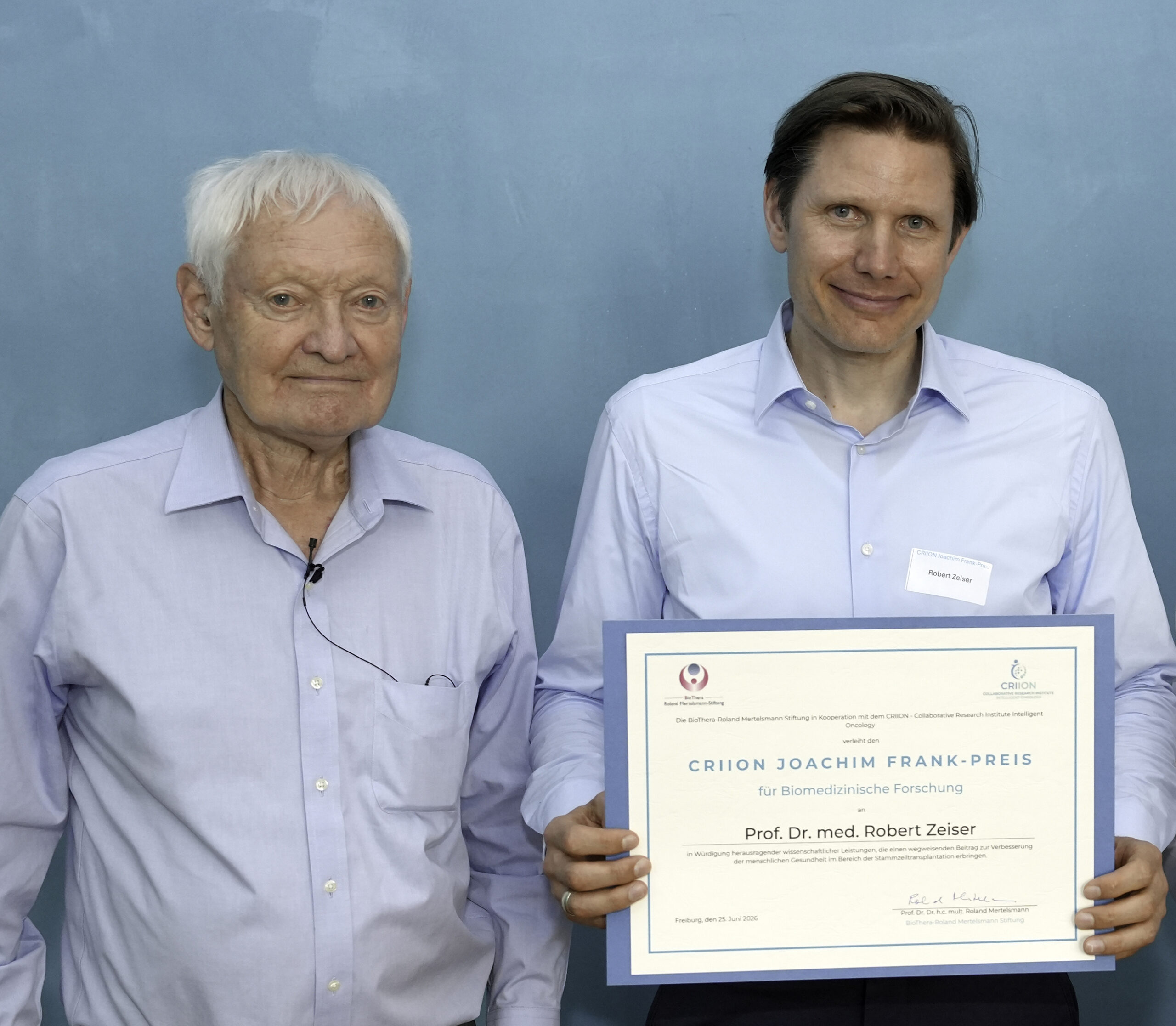 Two men in light blue shirts pose with a large framed certificate against a blue wall, suggesting an award ceremony.