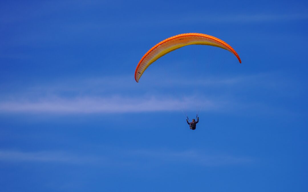 Paraglider with an orange-yellow wing gliding across a clear blue sky.