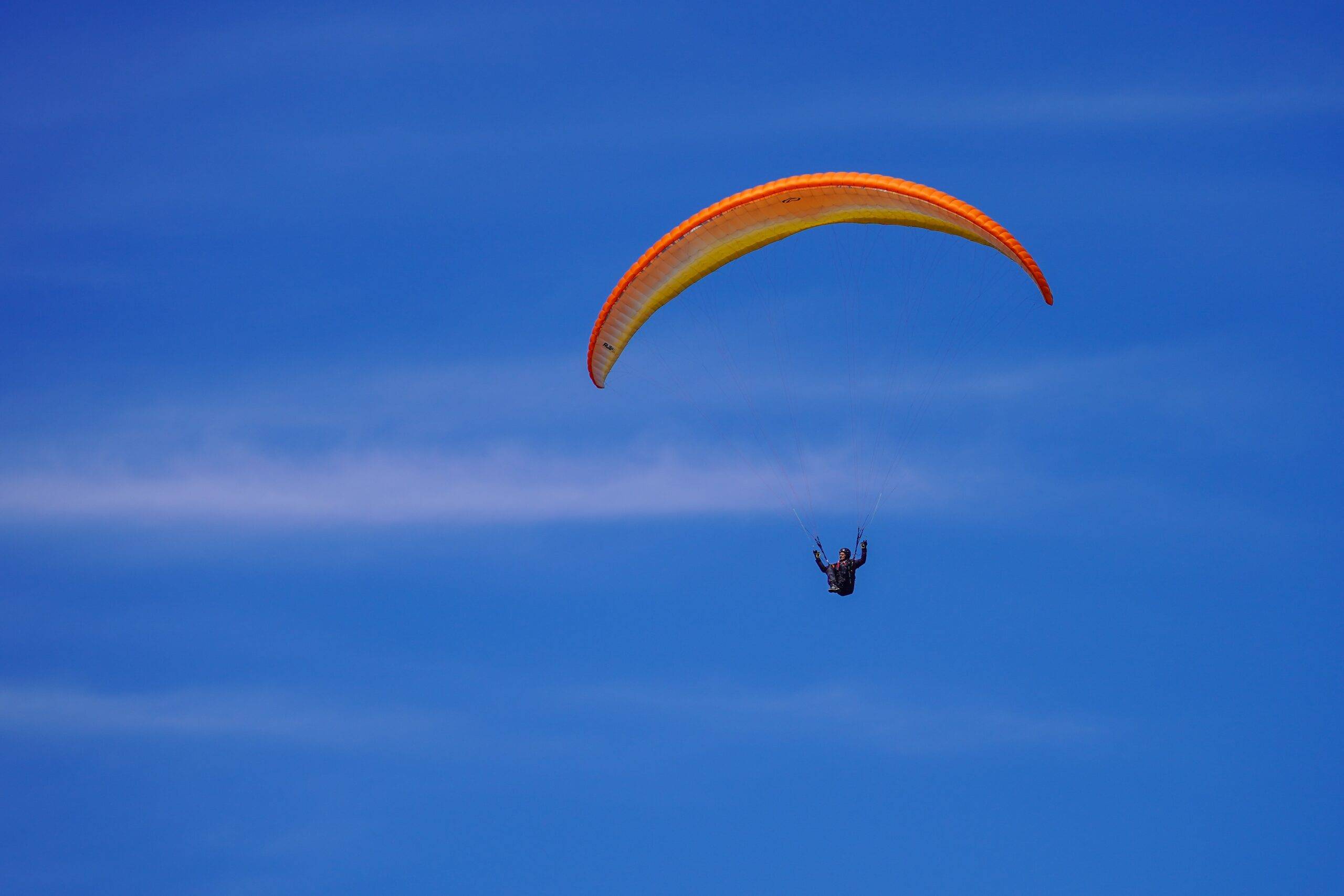Paraglider with an orange-yellow wing gliding across a clear blue sky.