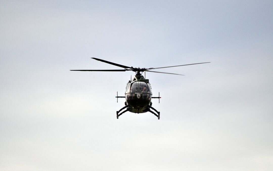 Front-on view of a helicopter in flight against a pale blue sky, rotor blades blurred from motion and landing skids visible beneath the fuselage.
