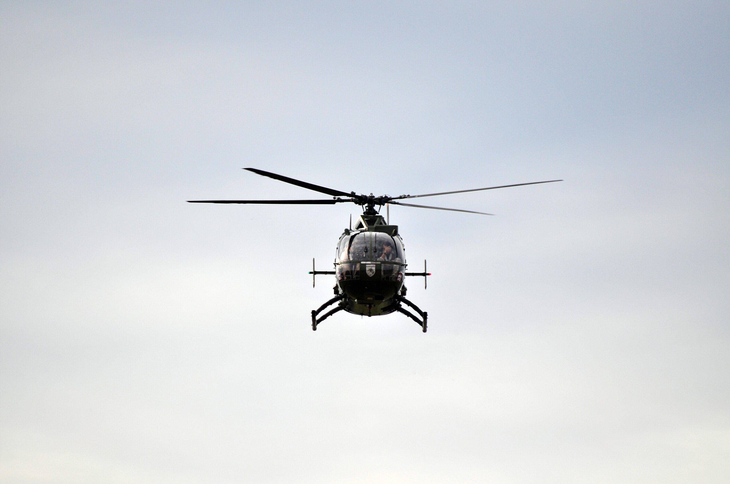 Front-on view of a helicopter in flight against a pale blue sky, rotor blades blurred from motion and landing skids visible beneath the fuselage.