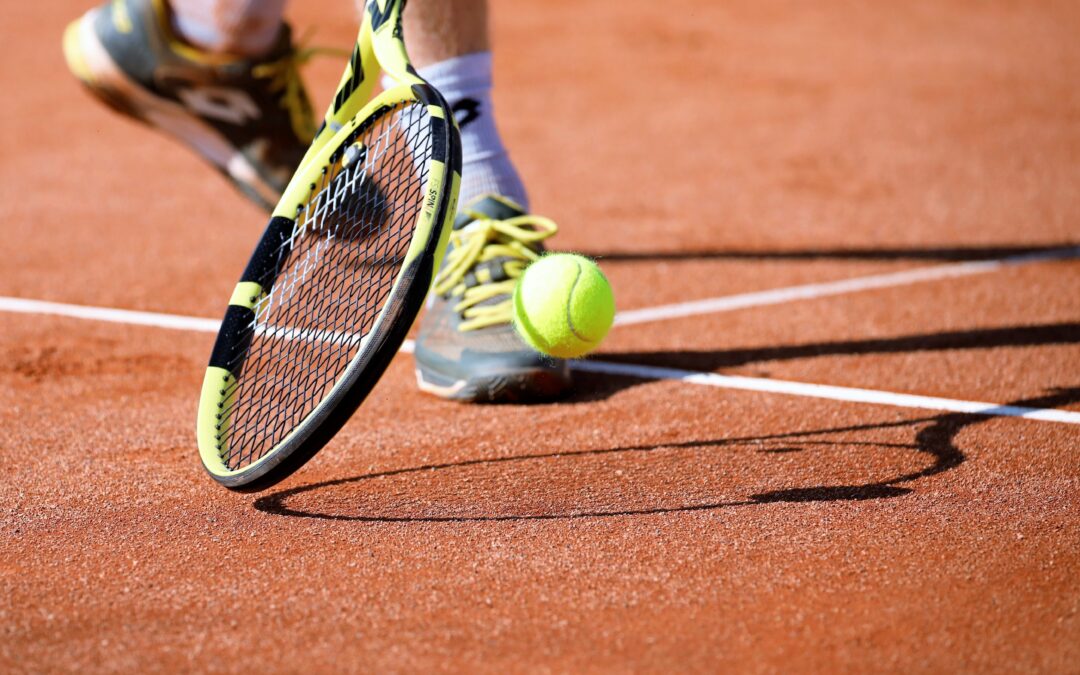 Close-up of a yellow-and-black tennis racket approaching a bright tennis ball on a red clay court.