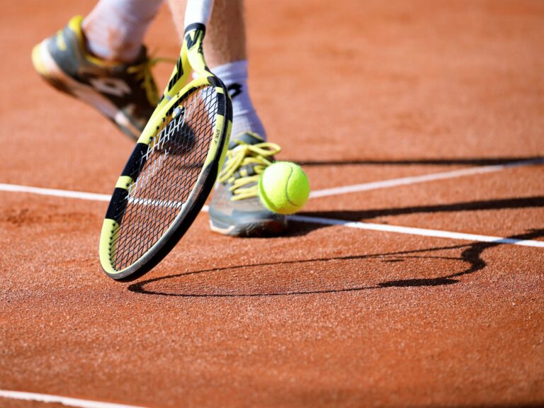 Close-up of a yellow-and-black tennis racket approaching a bright tennis ball on a red clay court.