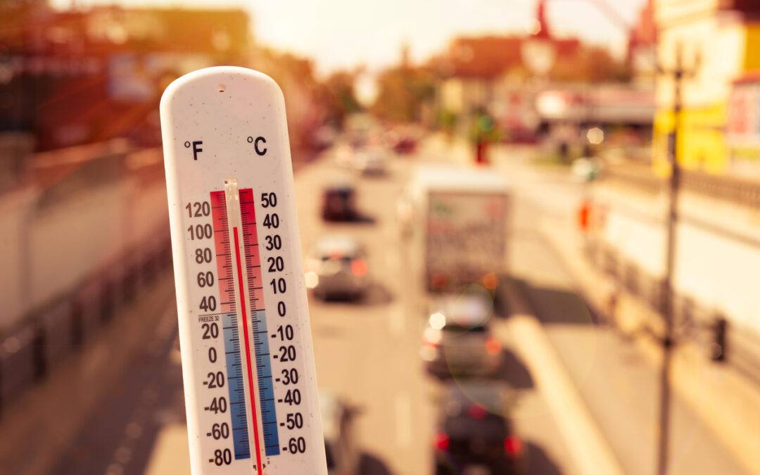 Close-up of an outdoor thermometer indicating warm weather, with a sunny urban street blur in the background.