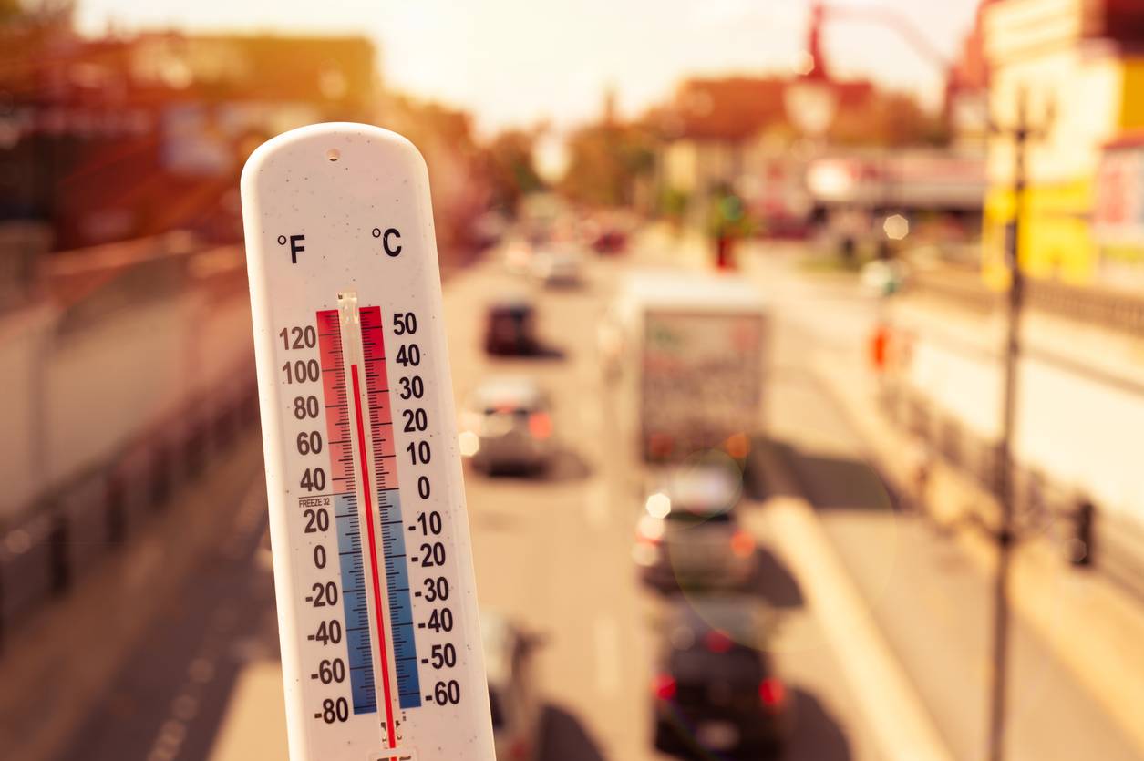 Close-up of an outdoor thermometer indicating warm weather, with a sunny urban street blur in the background.