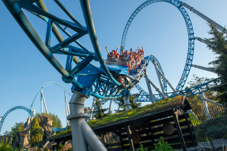 Riders raise hands in a blue roller coaster car as it curves through a steel track against a clear sky.