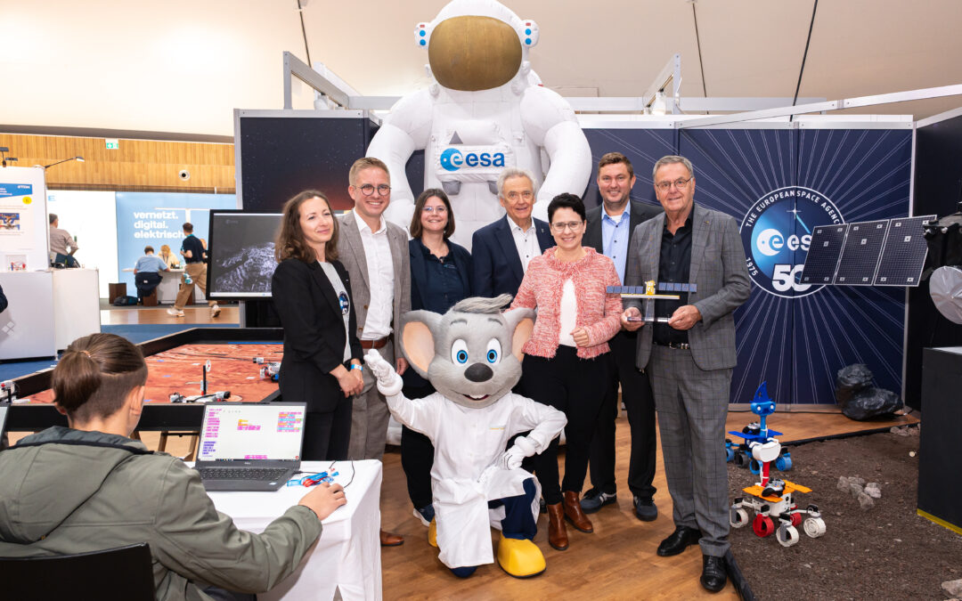Group of professionals posing with a large white ESA astronaut mascot at a science expo booth, smiling for the camera with laptops and robotics in view.