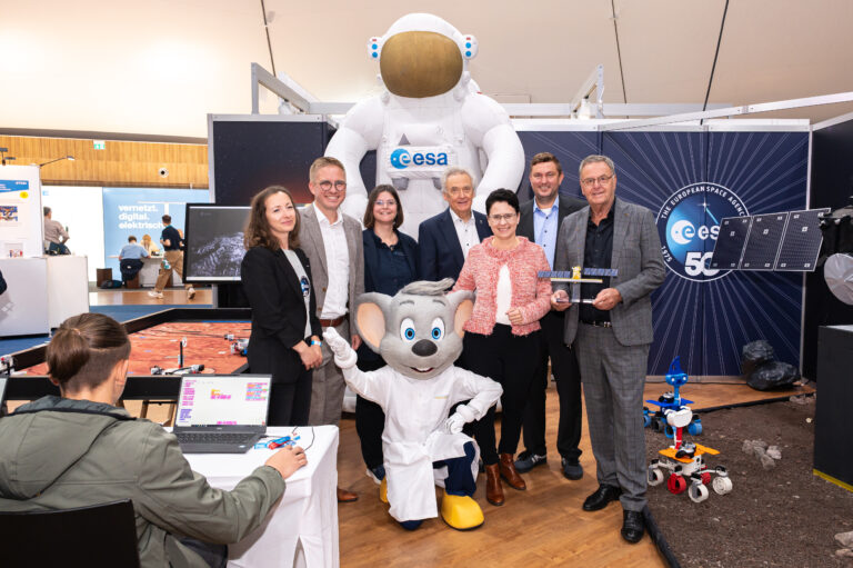 Group of professionals posing with a large white ESA astronaut mascot at a science expo booth, smiling for the camera with laptops and robotics in view.