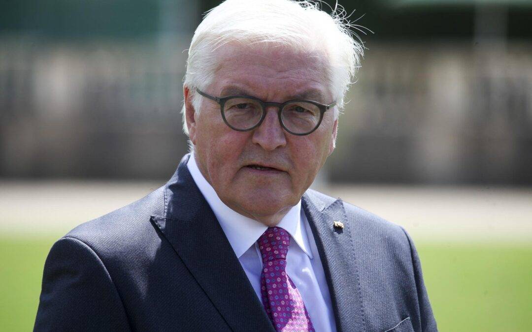 Older man in a navy suit, pink tie, and round glasses photographed outdoors.