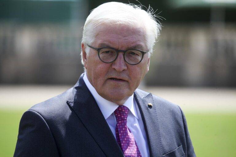 Older man in a navy suit, pink tie, and round glasses photographed outdoors.