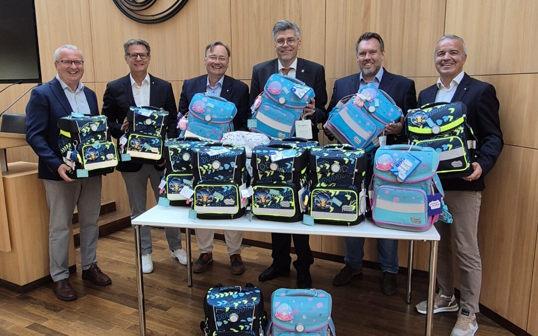 Six men in business attire pose with colorful backpacks in a wood-paneled conference room behind a table full of bags.