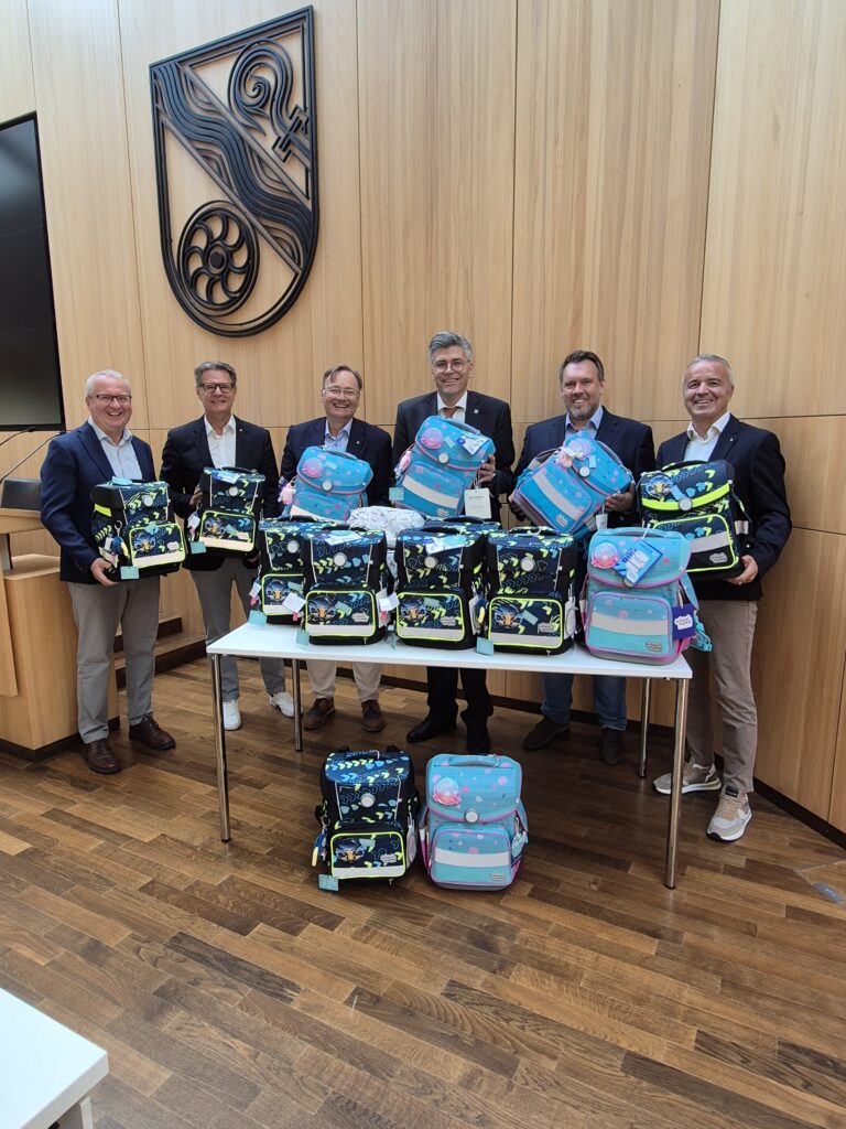 Six men in business attire pose with colorful backpacks in a wood-paneled conference room behind a table full of bags.