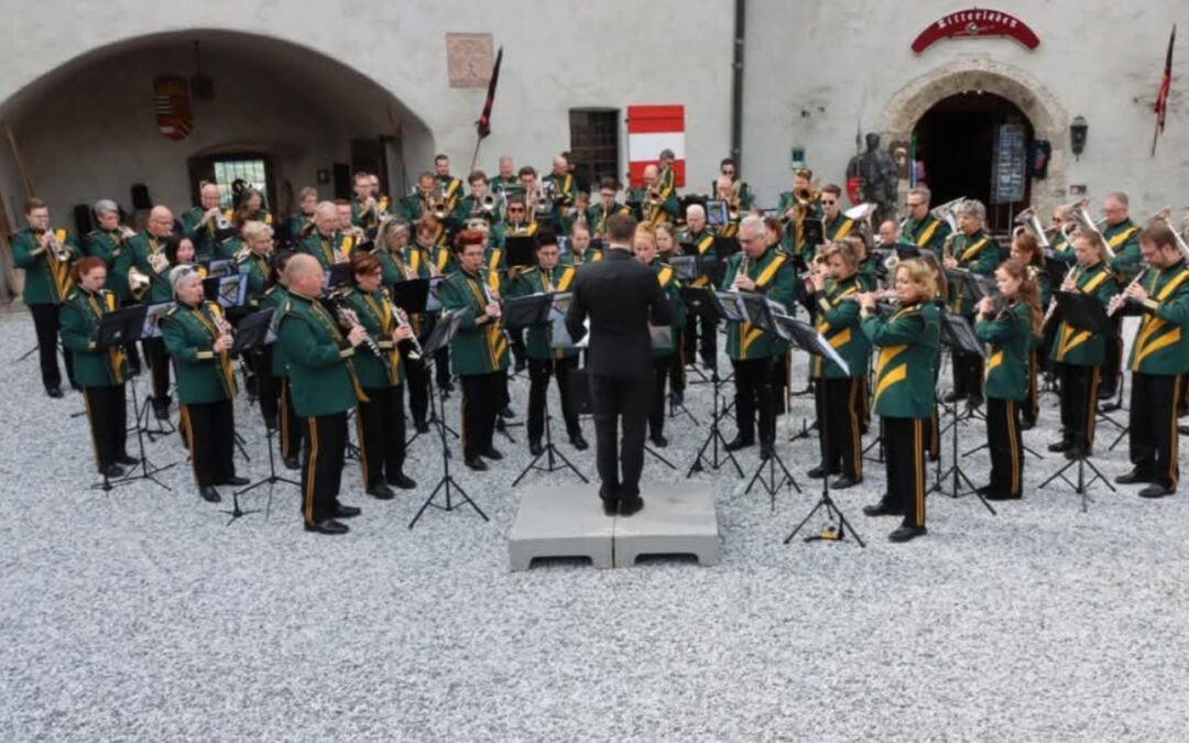 Large brass band performing outdoors in a historic courtyard, conductor on a small platform directing musicians in green uniforms with yellow sashes.