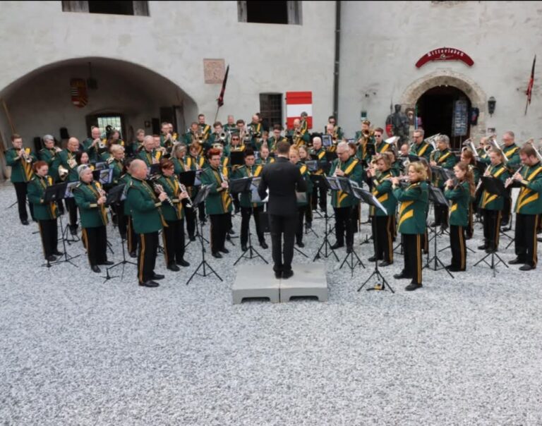 Large brass band performing outdoors in a historic courtyard, conductor on a small platform directing musicians in green uniforms with yellow sashes.