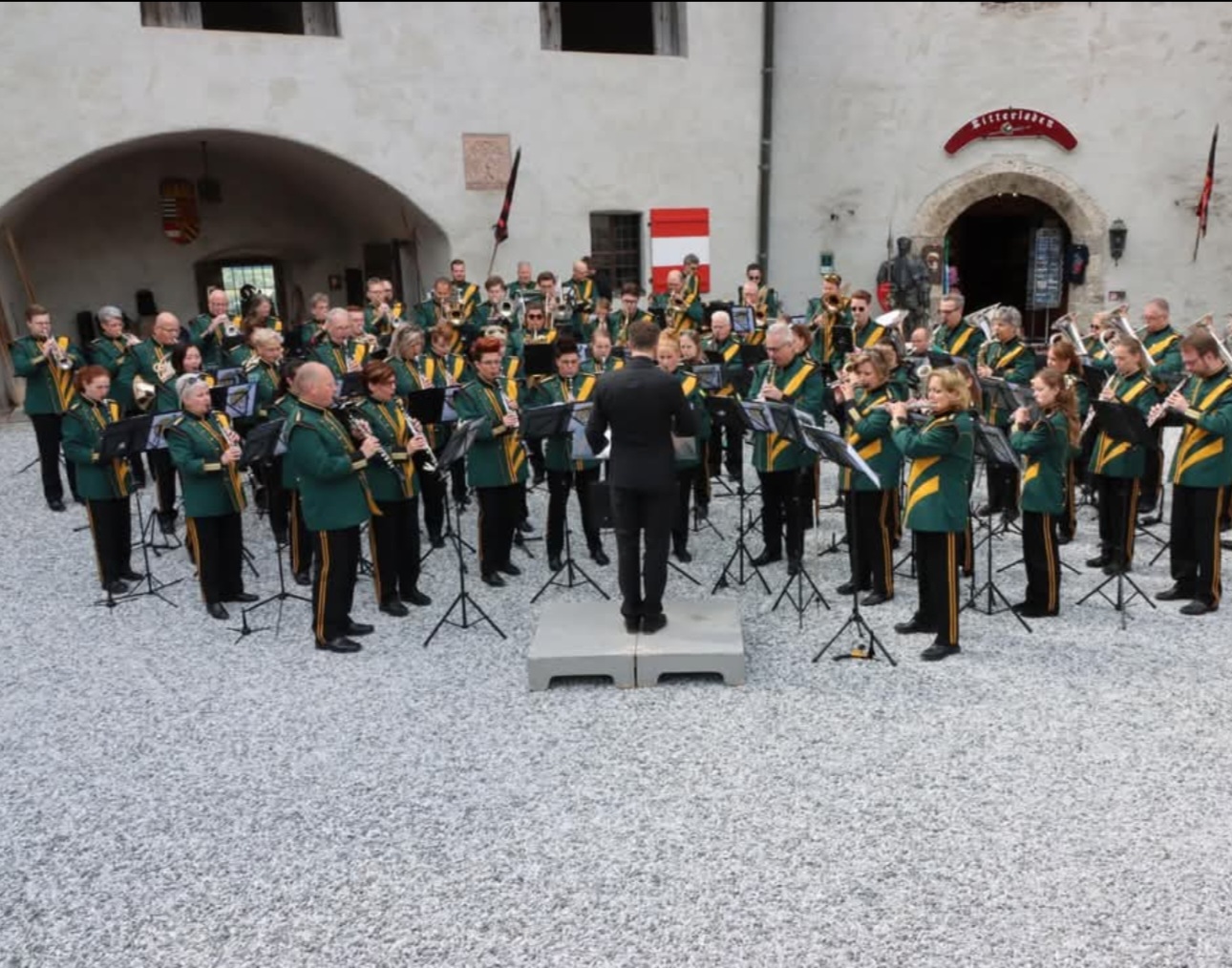 Large brass band performing outdoors in a historic courtyard, conductor on a small platform directing musicians in green uniforms with yellow sashes.