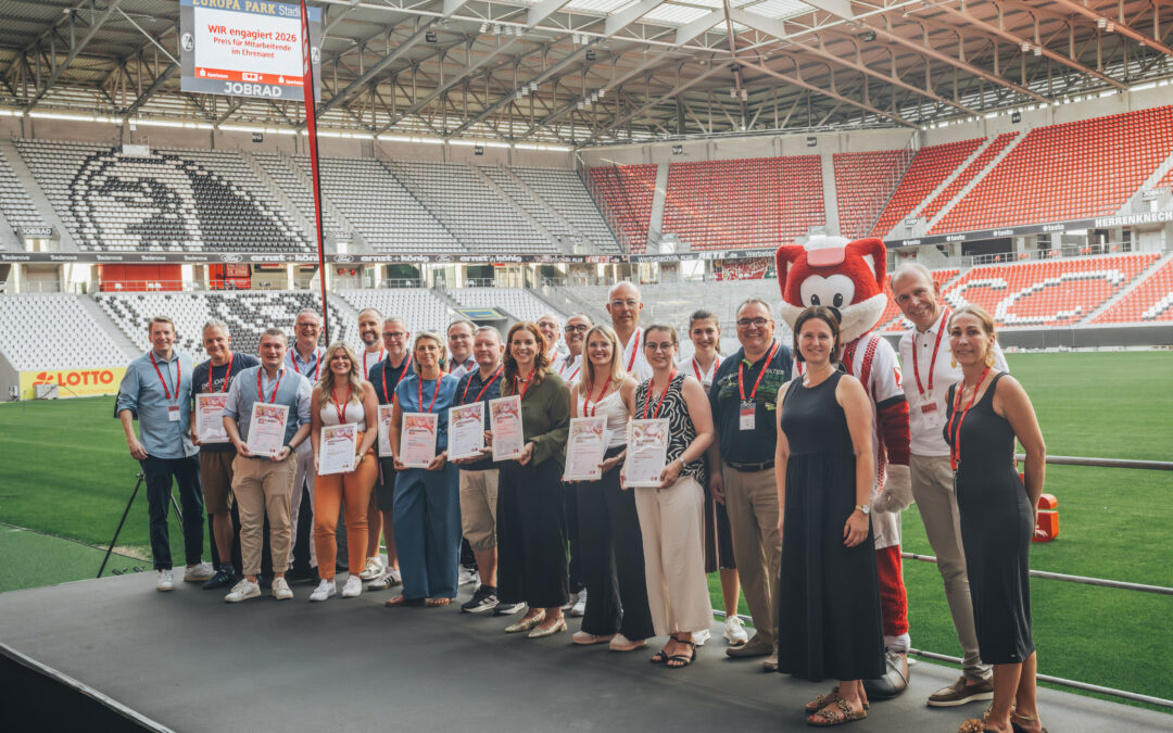 Group of people in a stadium posing with certificates, standing in a row beside a red mascot in a white outfit, on a green field under a large roof.