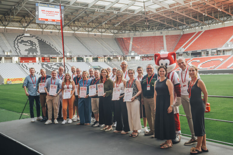 Group of people in a stadium posing with certificates, standing in a row beside a red mascot in a white outfit, on a green field under a large roof.