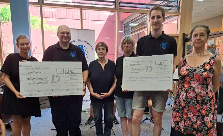 Six people indoors, two holding oversized donation checks and smiling for the photo.