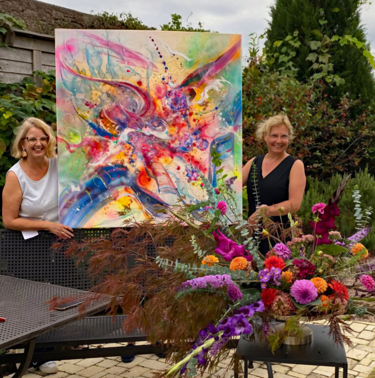 Outdoor display of a large colorful abstract painting on an easel, with two smiling women nearby and vibrant flowers in the foreground and background.