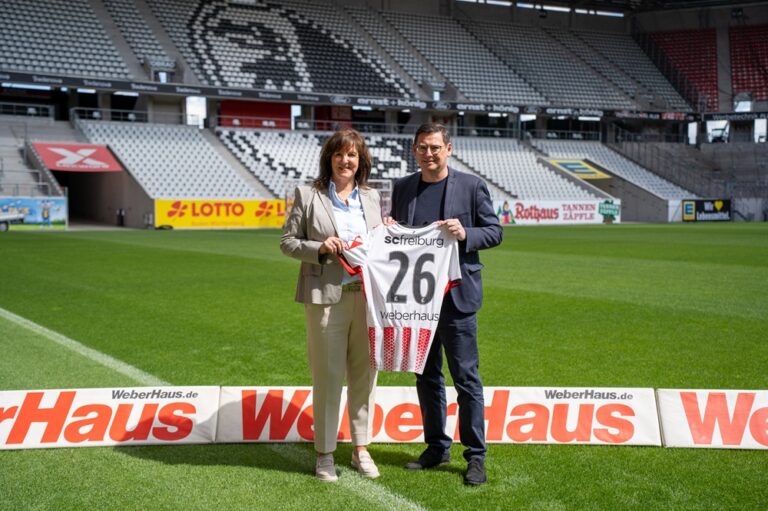 Two people stand on a football pitch, holding a white jersey with the number 26, with empty stands in the background and ads along the sideline.
