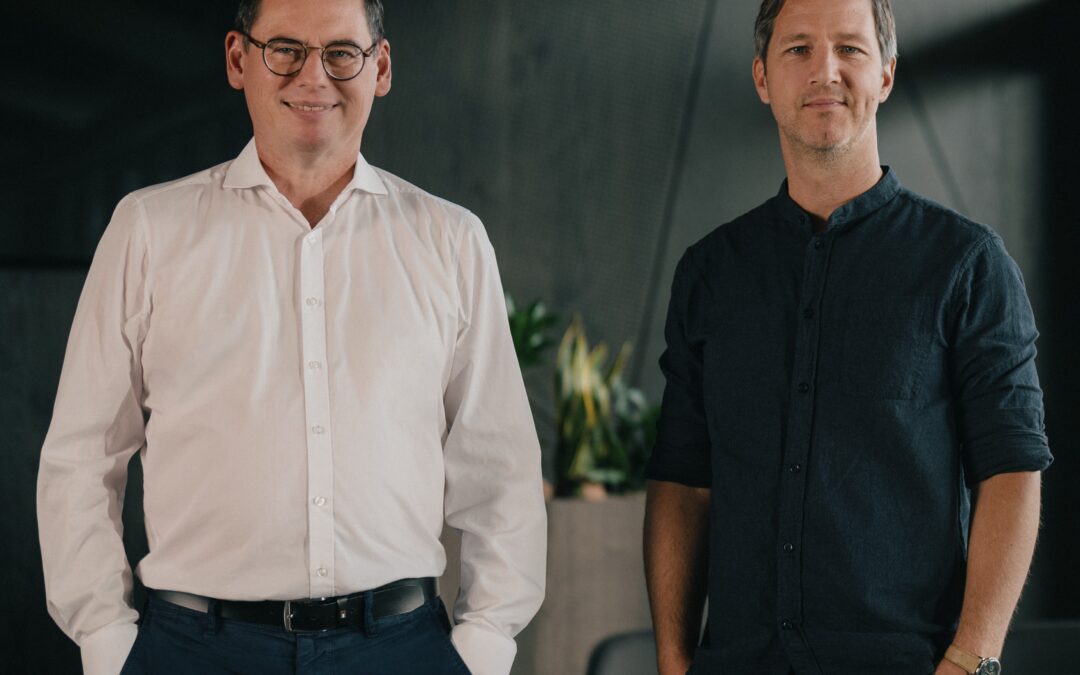 Two men stand side by side in a modern office, one in a white shirt with glasses and the other in a dark shirt, both smiling subtly.