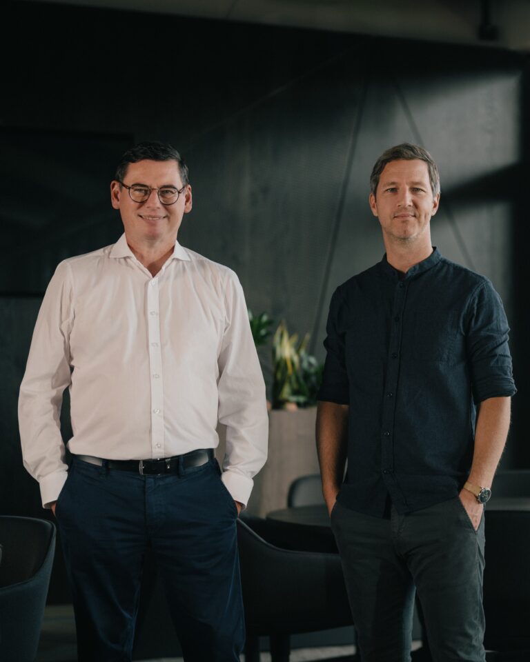 Two men stand side by side in a modern office, one in a white shirt with glasses and the other in a dark shirt, both smiling subtly.