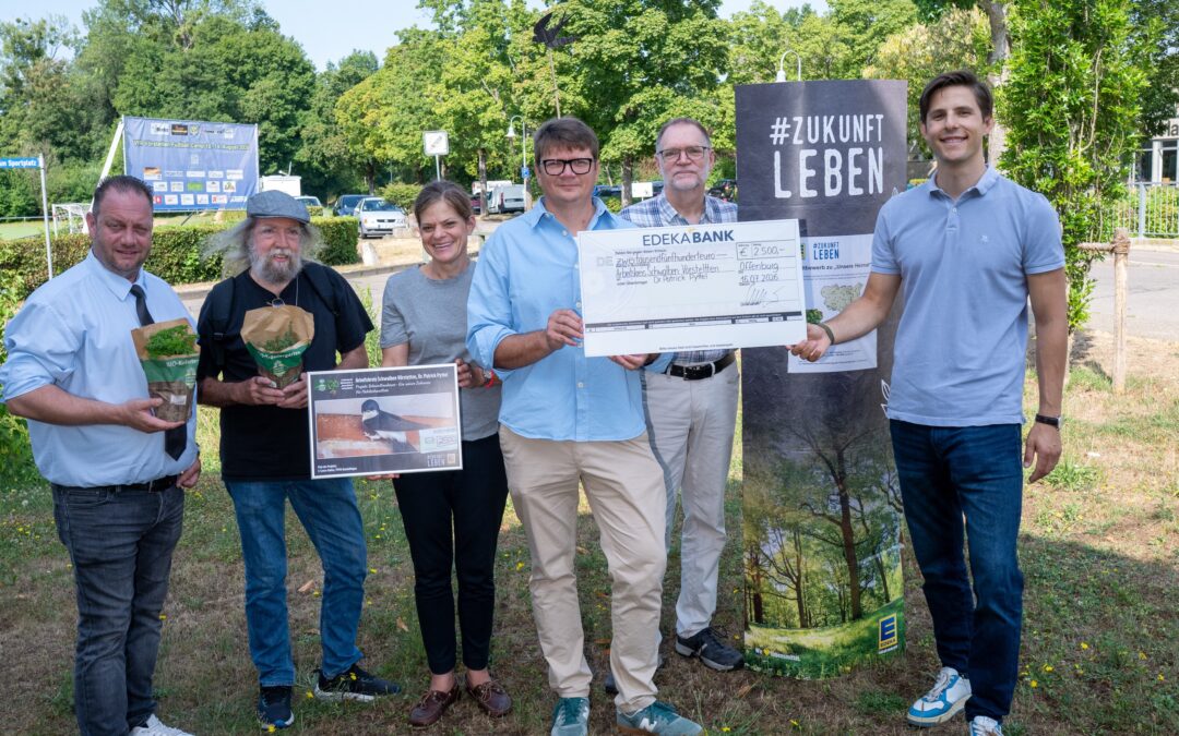 Six adults stand outdoors, smiling, holding a large ceremonial check and event posters with a '#ZUKUNFT LEBEN' banner visible in the background.
