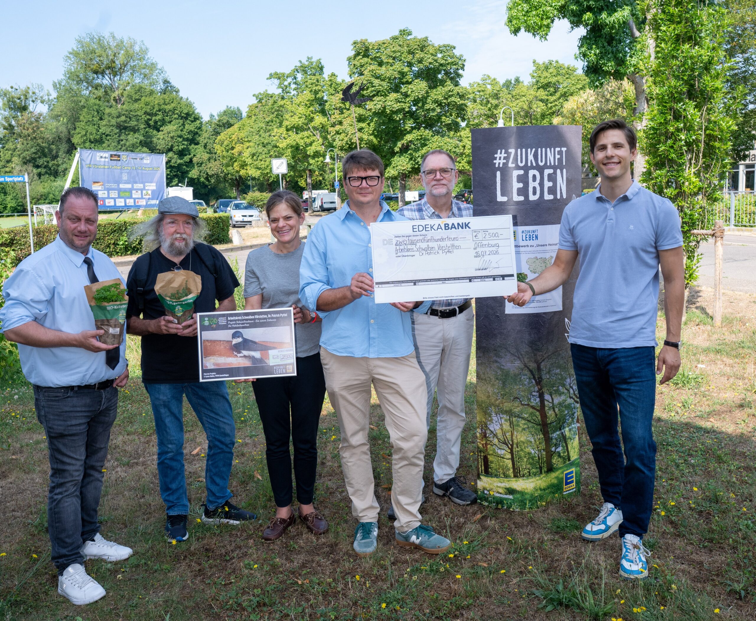 Six adults stand outdoors, smiling, holding a large ceremonial check and event posters with a '#ZUKUNFT LEBEN' banner visible in the background.