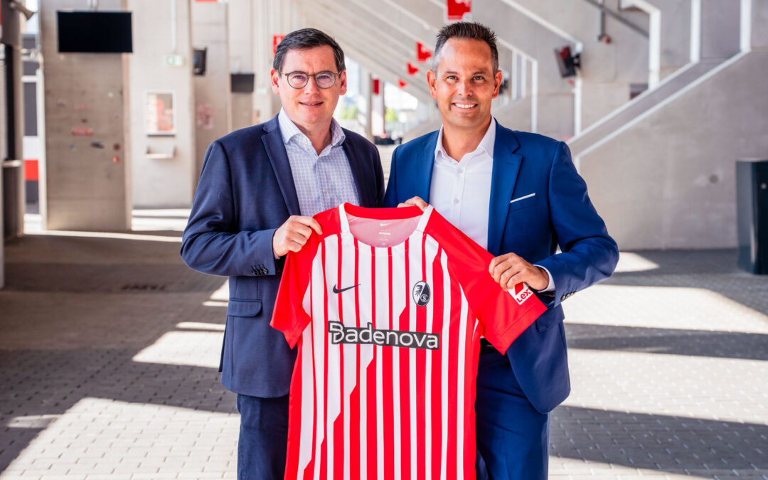 Two men in suits smile as they hold up a red-and-white striped soccer jersey in a bright, modern space.