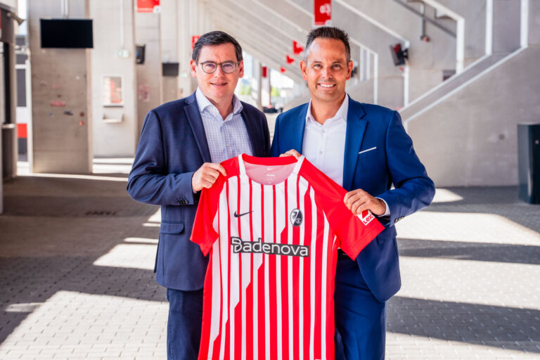 Two men in suits smile as they hold up a red-and-white striped soccer jersey in a bright, modern space.