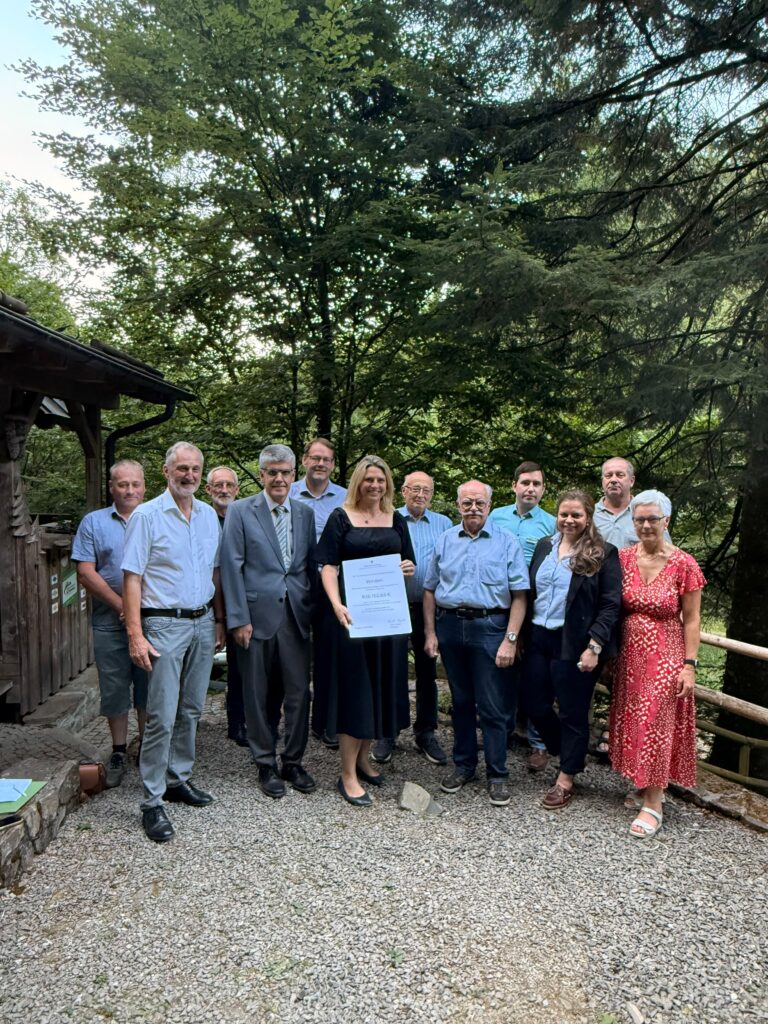 Group of adults posing outdoors on a gravel path, trees behind them; one woman in black dress holds a large document or certificate while others smile in business casual attire.