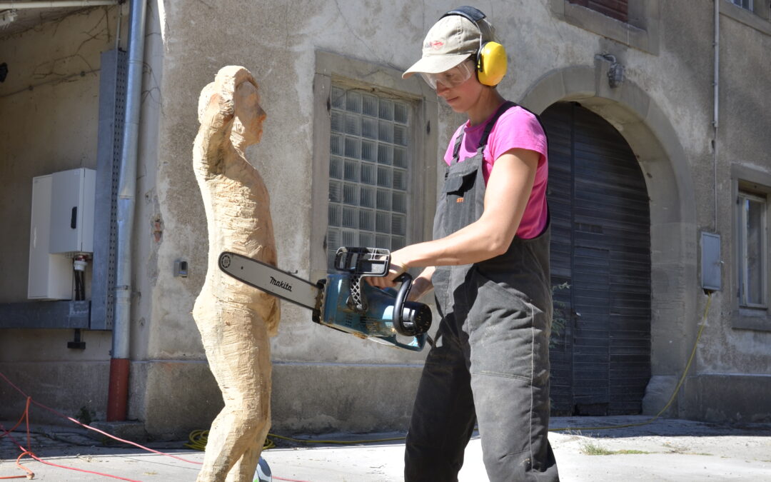 Worker wearing pink shirt and overalls with yellow ear protection uses a power saw to carve a wooden sculpture outdoors.