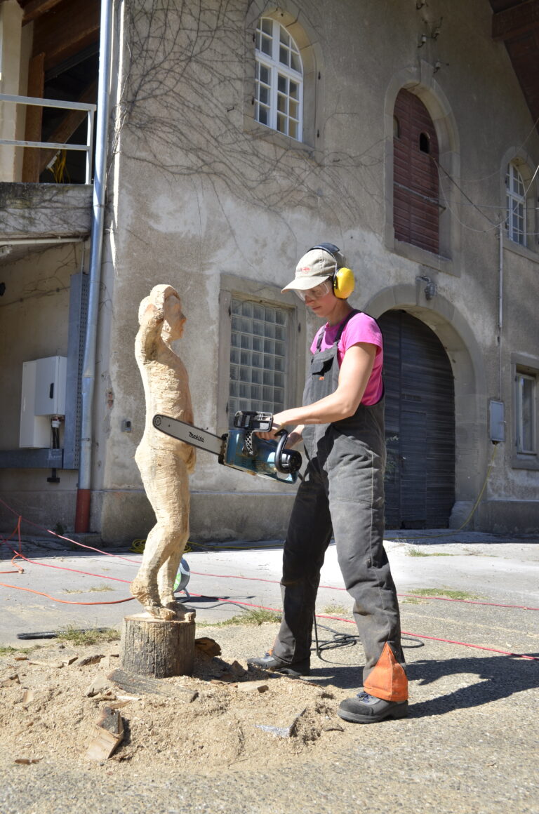 Worker wearing pink shirt and overalls with yellow ear protection uses a power saw to carve a wooden sculpture outdoors.