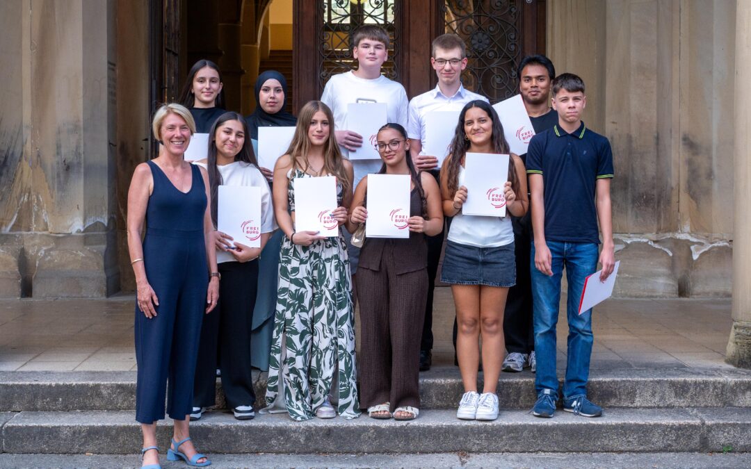 Group of nine students and a woman teacher standing on stone steps outside a building, each student holding a white certificate or folder with a red circular logo.