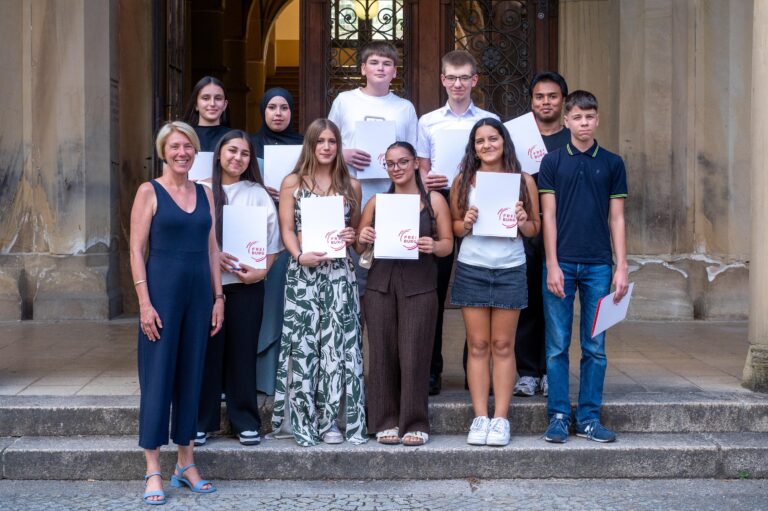 Group of nine students and a woman teacher standing on stone steps outside a building, each student holding a white certificate or folder with a red circular logo.
