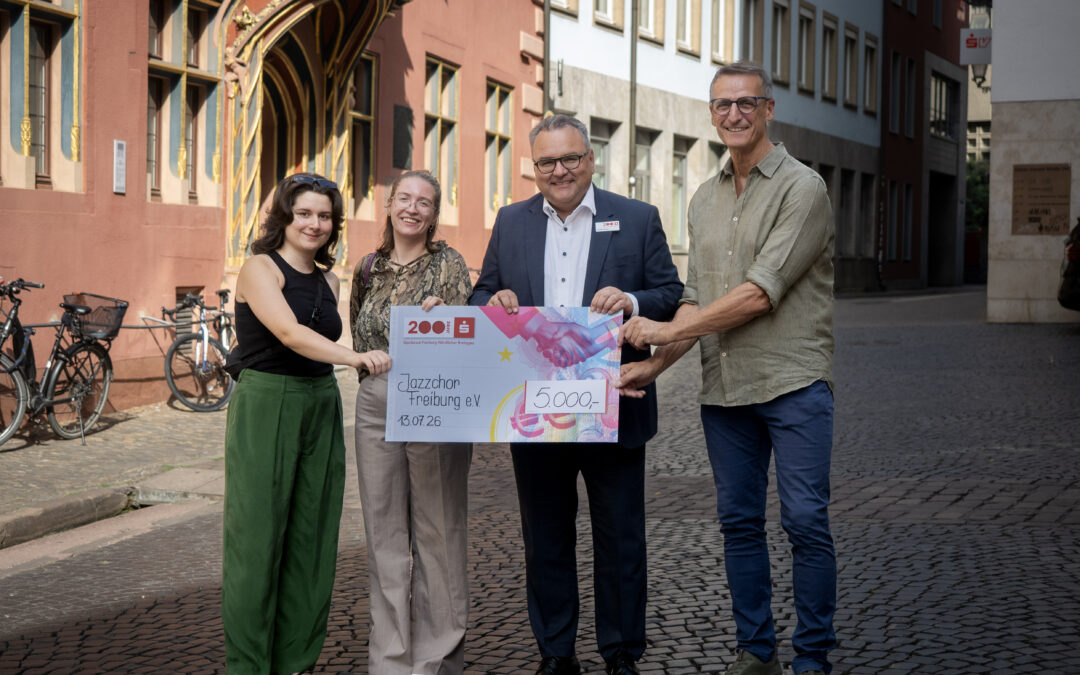 Four people on a cobblestone street present a large, colorful donation check, smiling for the photo in front of historic buildings.