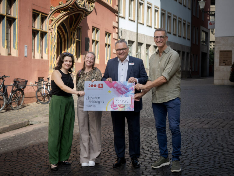 Four people on a cobblestone street present a large, colorful donation check, smiling for the photo in front of historic buildings.