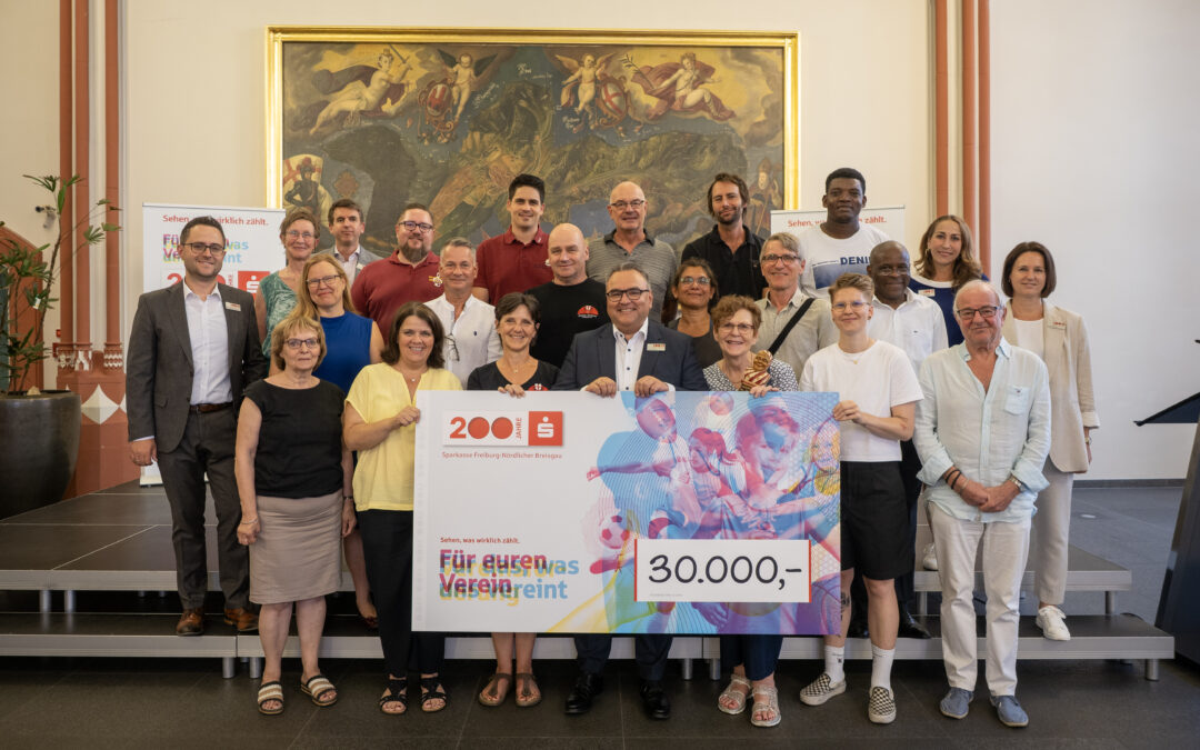 Group of diverse adults posing on steps indoors, holding a large colorful cheque for 30,000 euros and celebrating a donation event.