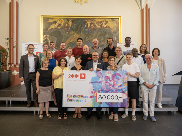 Group of diverse adults posing on steps indoors, holding a large colorful cheque for 30,000 euros and celebrating a donation event.