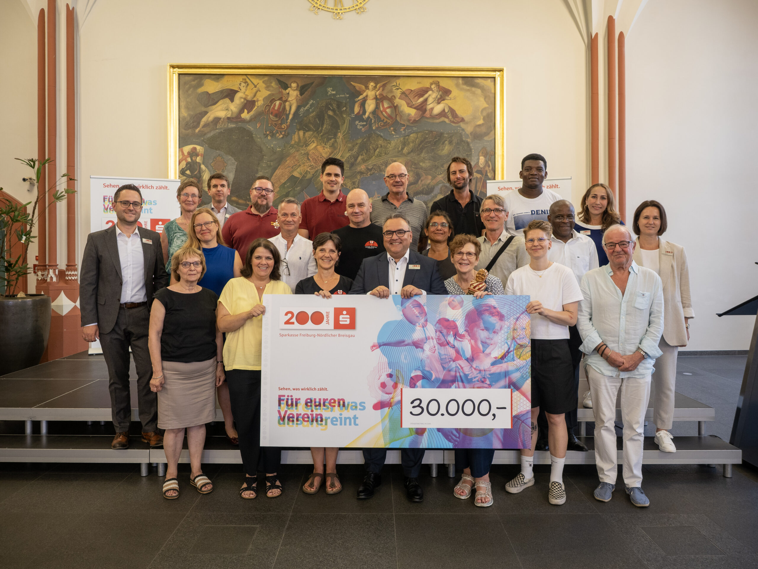 Group of diverse adults posing on steps indoors, holding a large colorful cheque for 30,000 euros and celebrating a donation event.