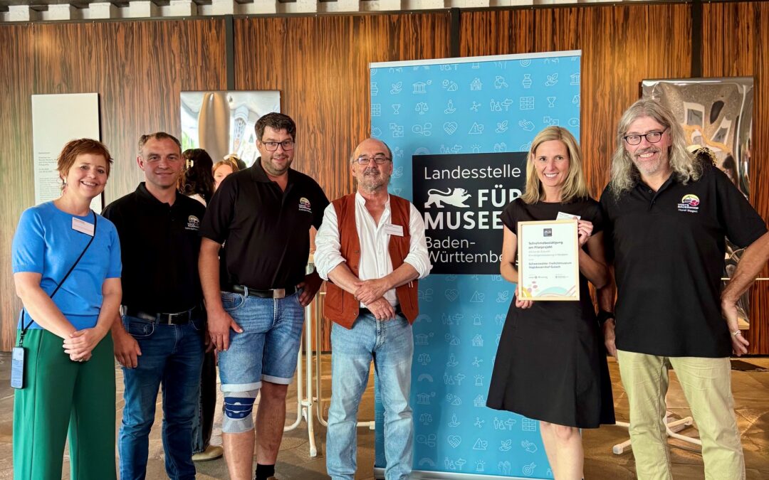 Six adults standing indoors, posing beside a blue banner that reads 'Landesstelle Für Museum...' One woman holds a framed certificate; others wear name badges and casual/work attire, with a wooden wall backdrop.