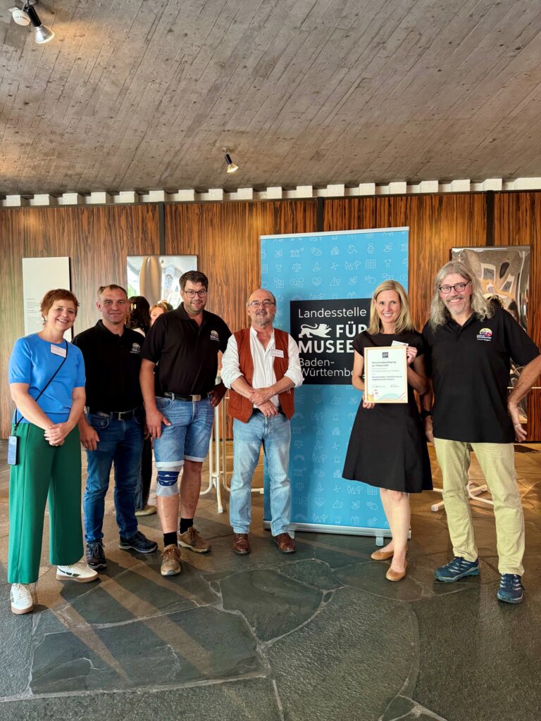 Six adults standing indoors, posing beside a blue banner that reads 'Landesstelle Für Museum...' One woman holds a framed certificate; others wear name badges and casual/work attire, with a wooden wall backdrop.