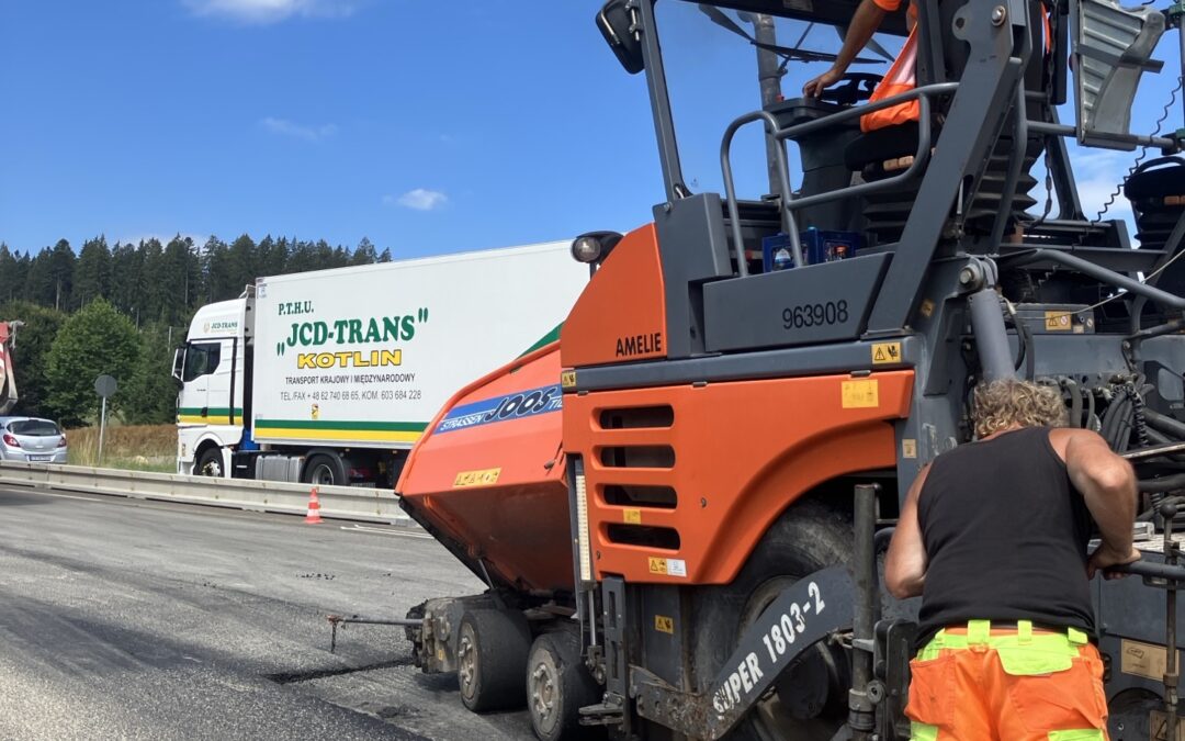 Construction workers in orange safety gear operate a large orange road roller on a highway under a blue sky with a white truck in the background