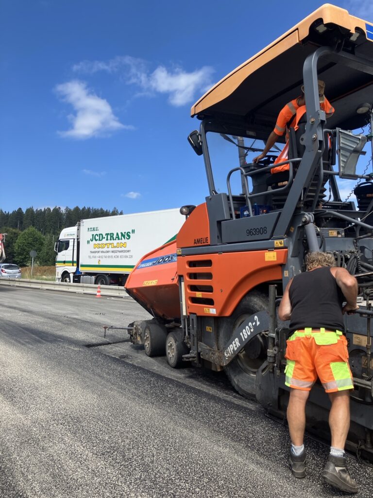Construction workers in orange safety gear operate a large orange road roller on a highway under a blue sky with a white truck in the background