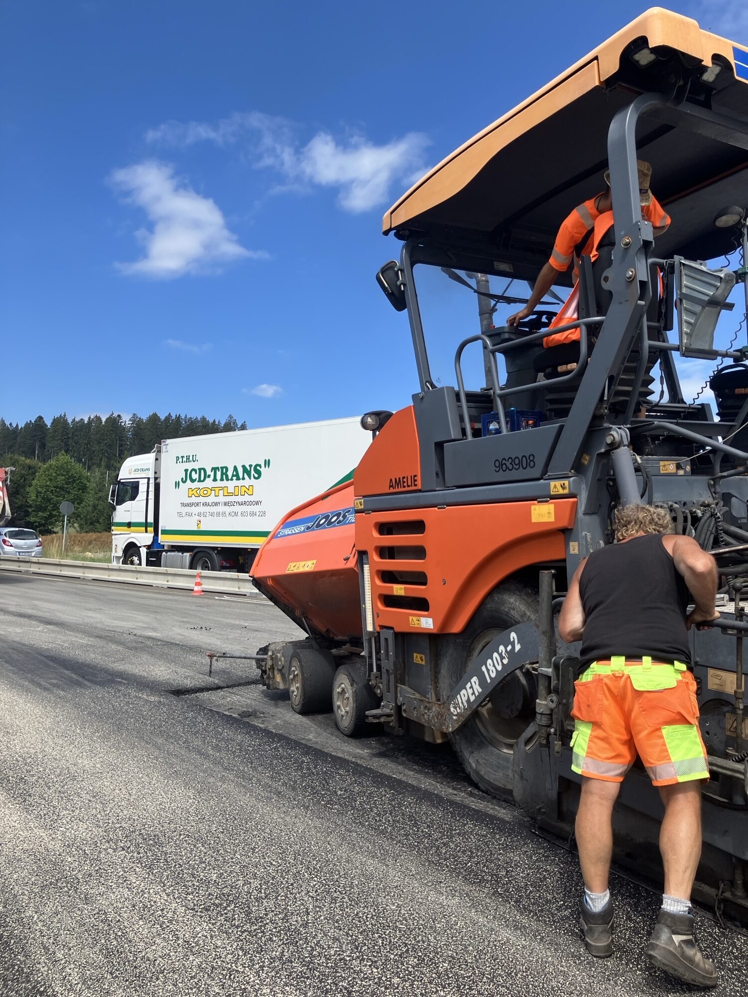 Construction workers in orange safety gear operate a large orange road roller on a highway under a blue sky with a white truck in the background